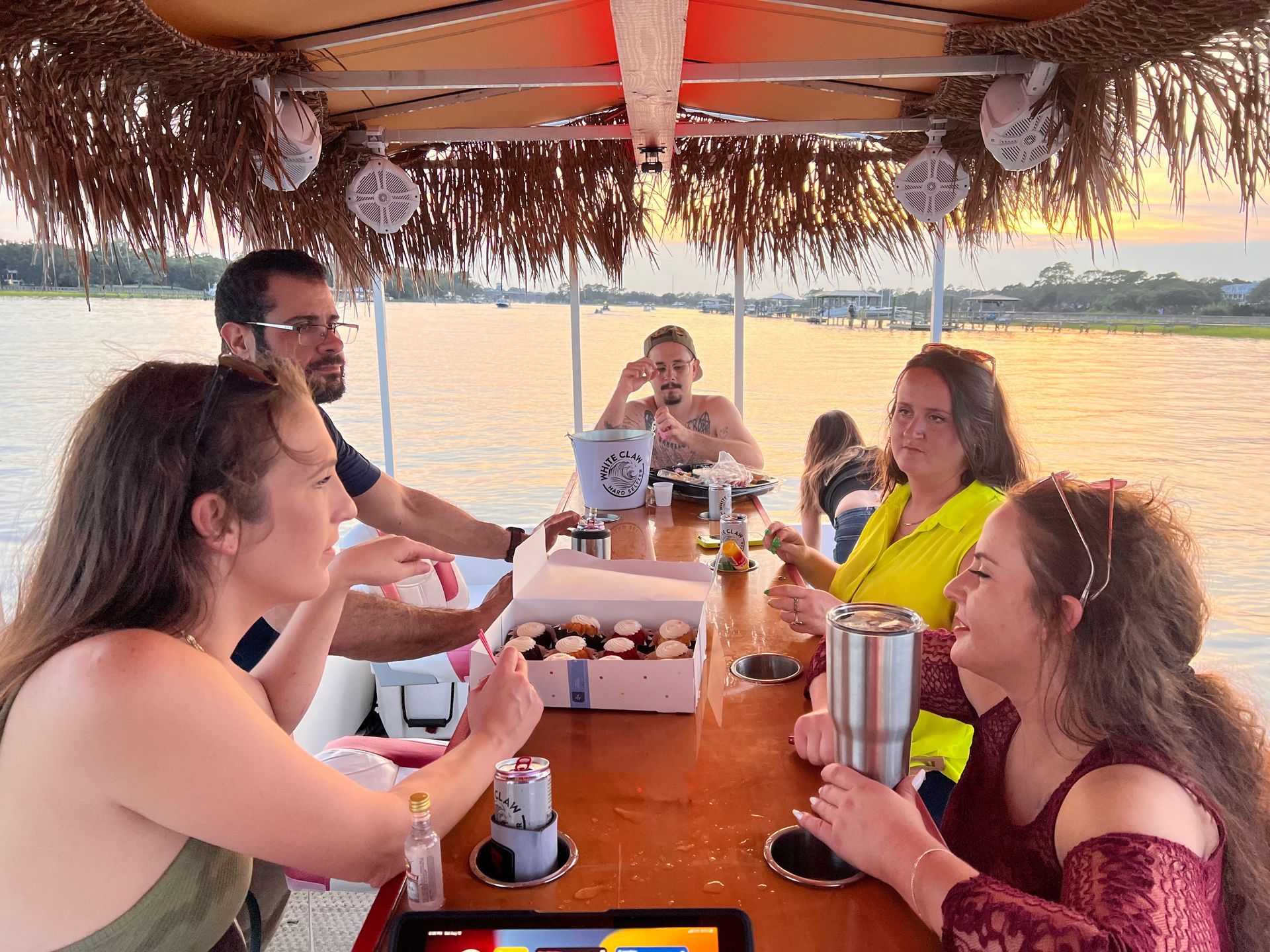 Friends seated at a wooden table in a thatched riverside boat, sharing drinks at sunset