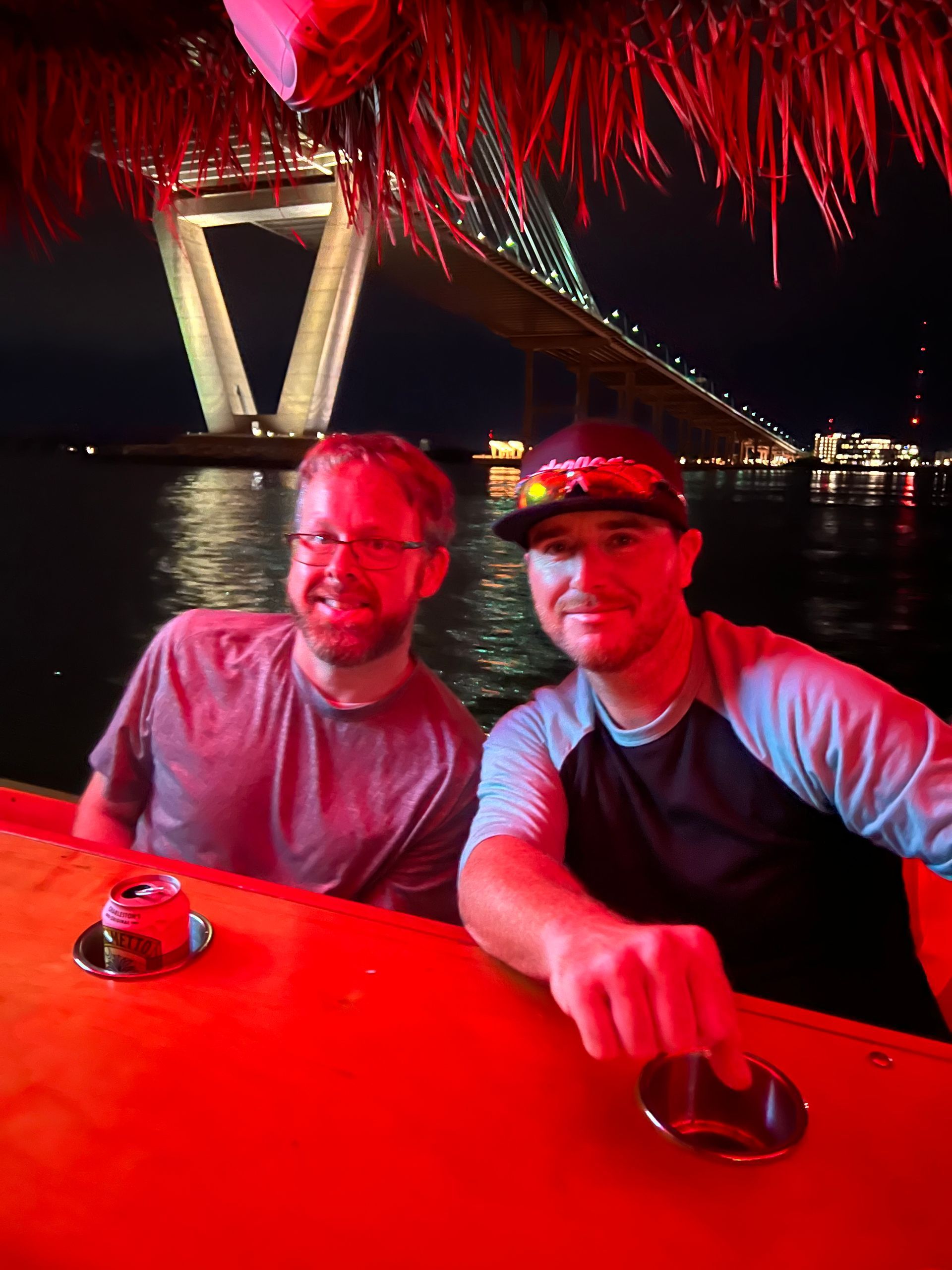 Two men on a boat at night under a lit bridge, with red lighting and water in the background.