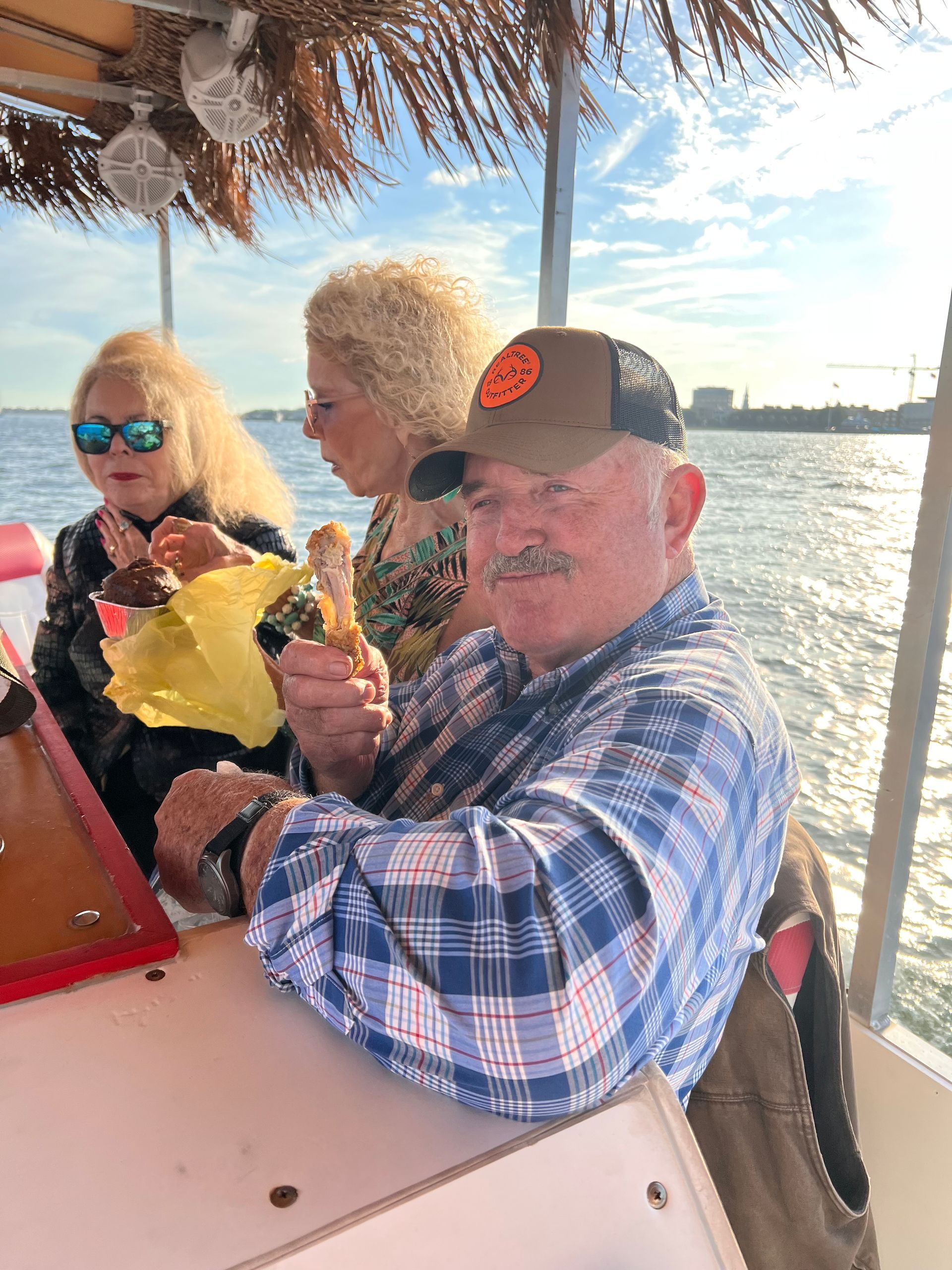 Two people at a tiki boat table holding drinks, with water and a city skyline in the background.