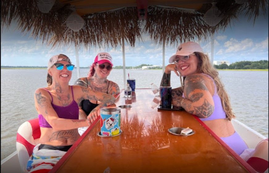 Three people smiling at a wooden table on a boat, with a lake and cloudy sky in the background