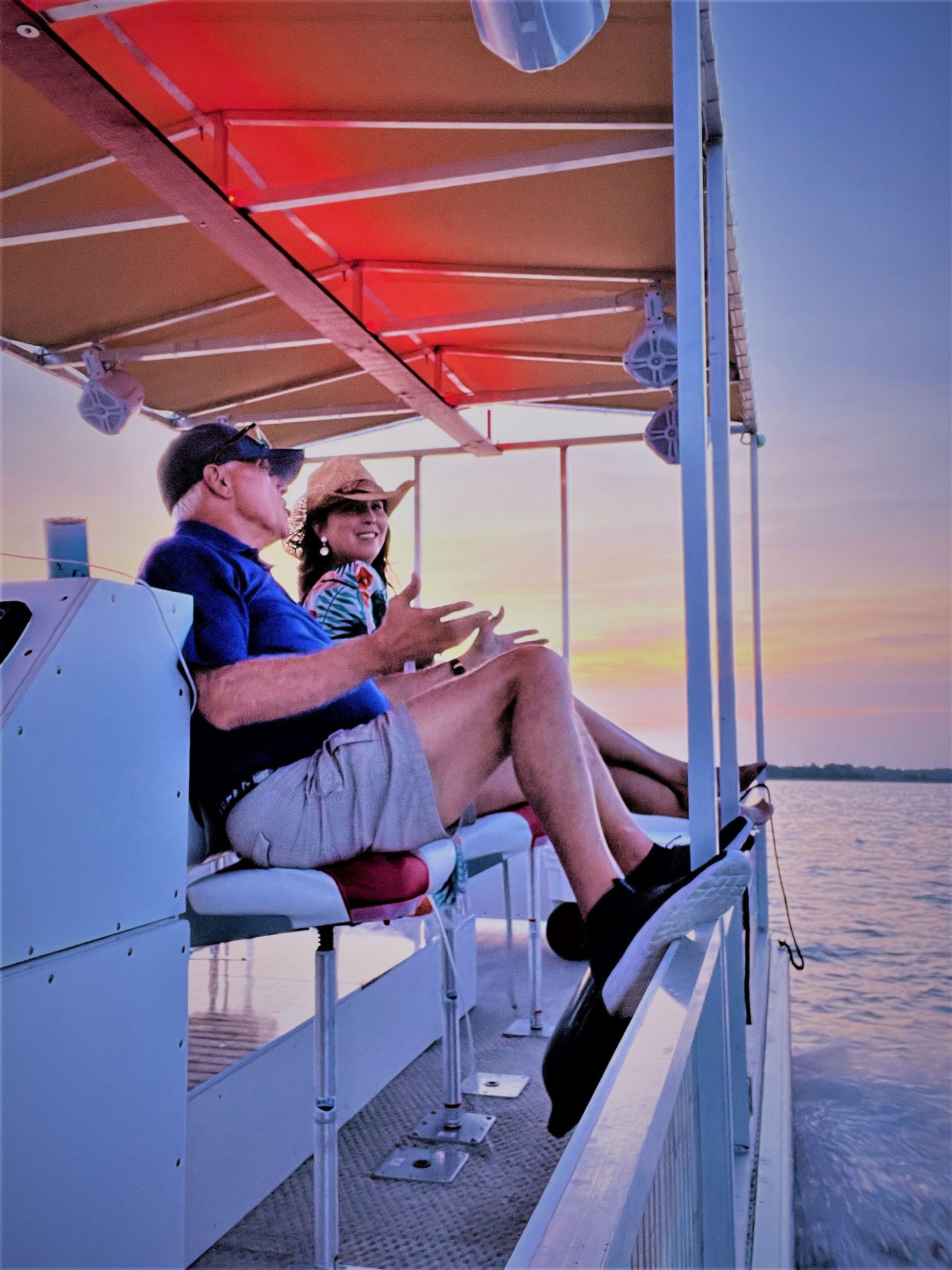 Two people sit on a boat under a red canopy at sunset, looking out over calm water.