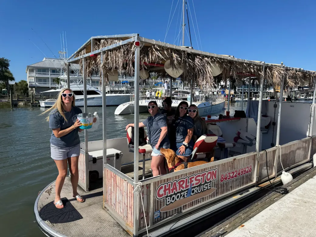 People on a decorated boat at a marina, waving near a dock under a blue sky.