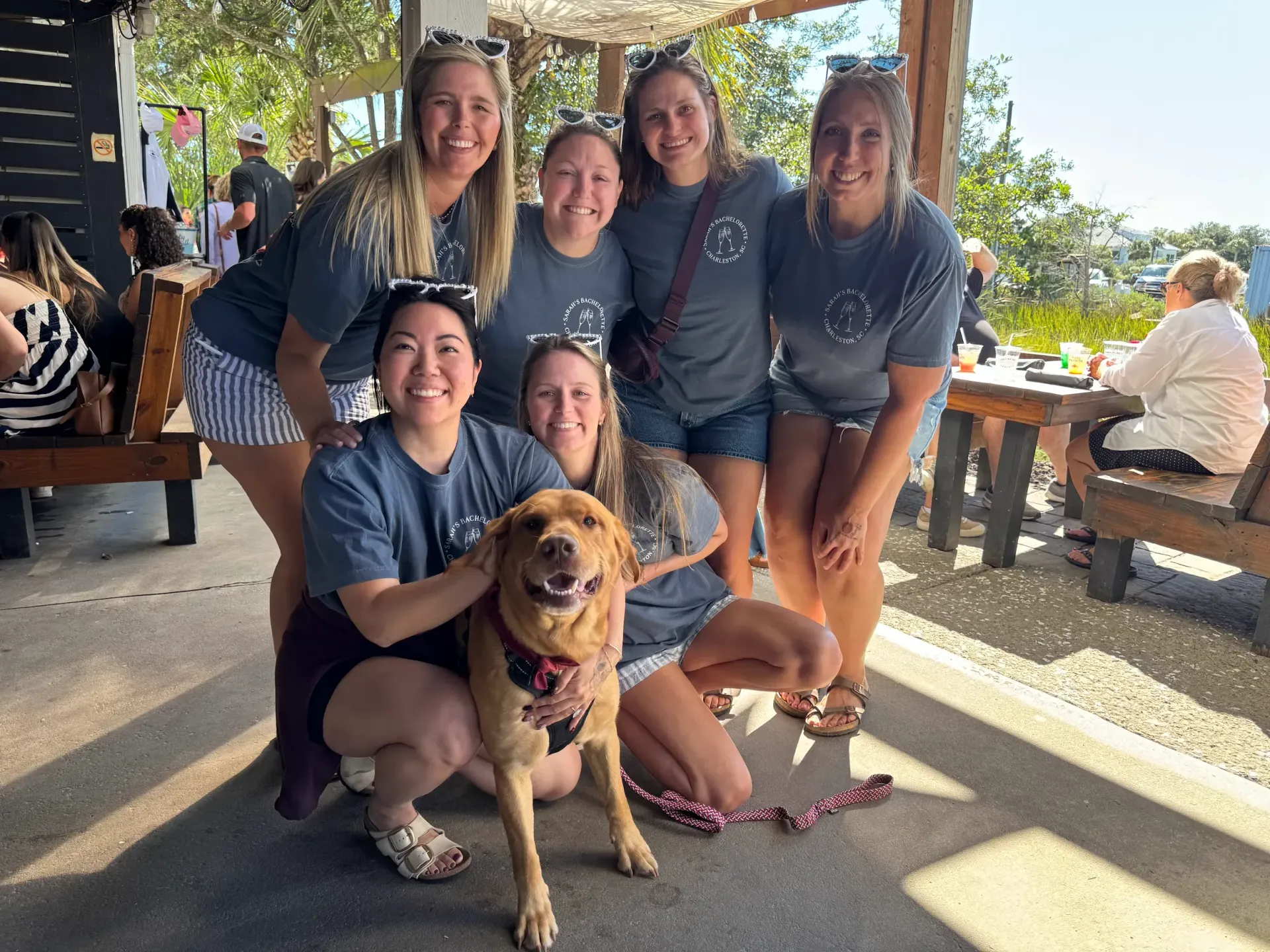 Five people pose with a dog at an outdoor café, smiling in the shade.