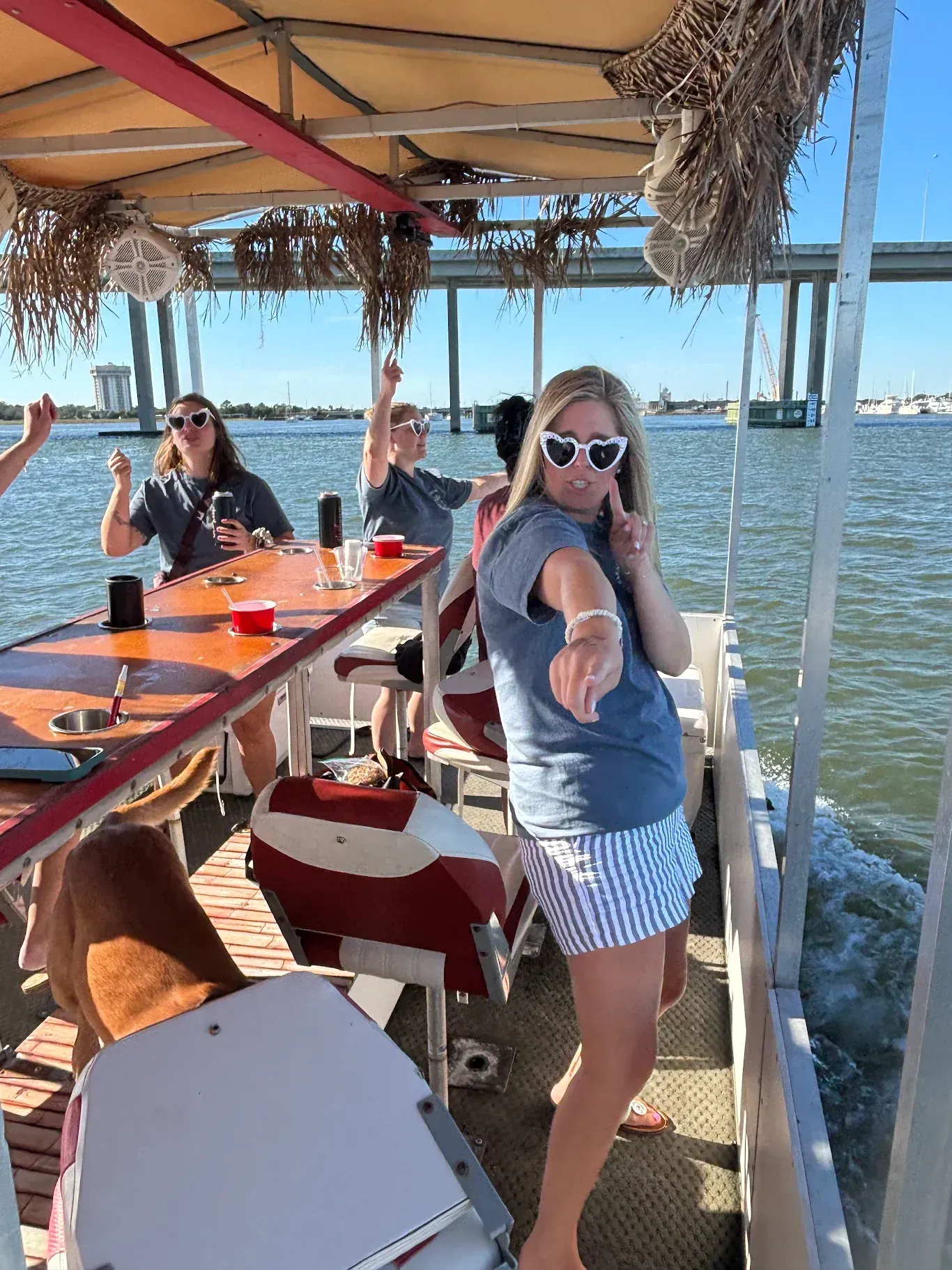Woman in sunglasses on a covered boat deck, pointing toward the camera beside a table on the water.