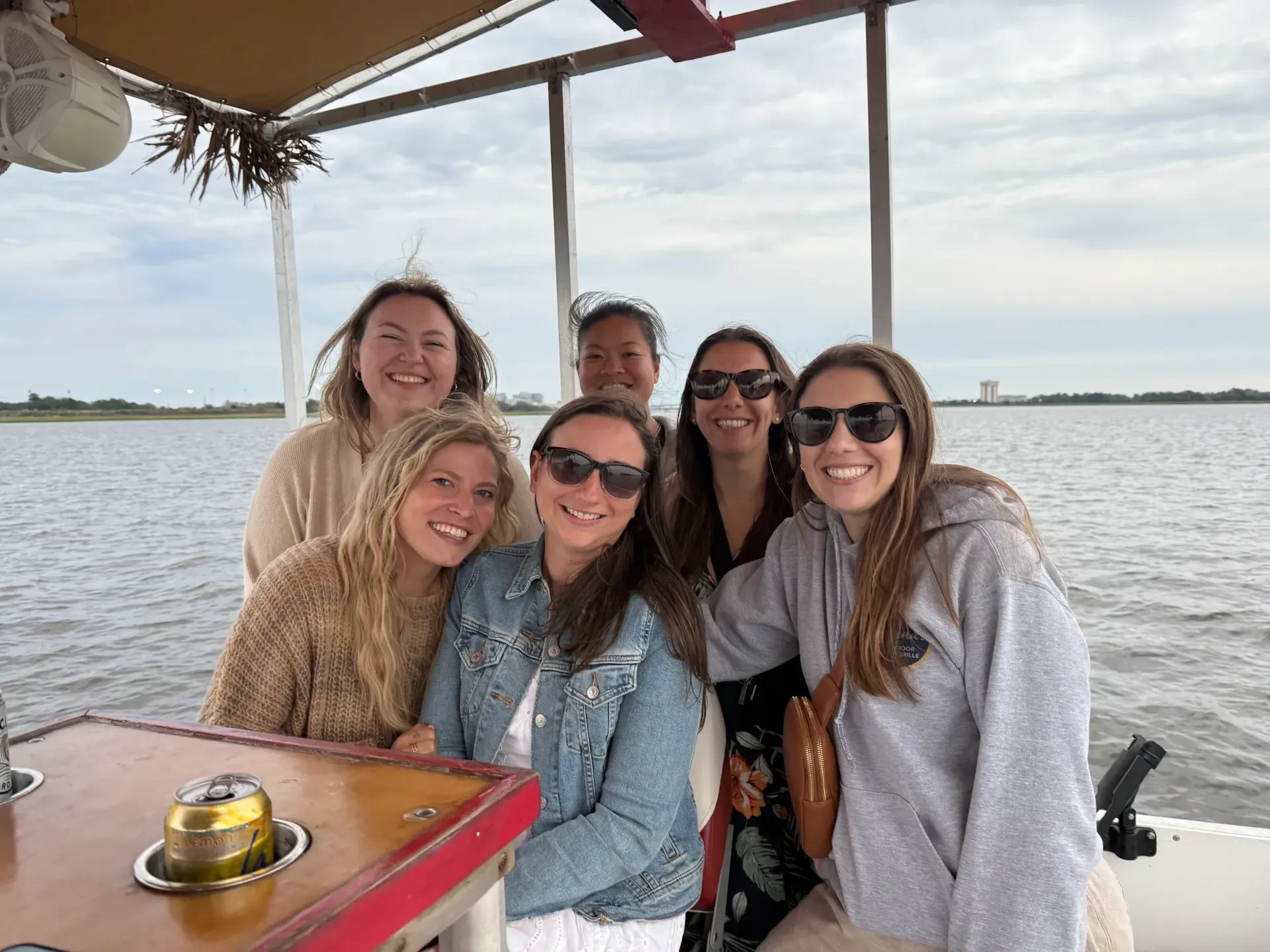 Five friends smiling on a boat, seated at a table on calm water under a canopy