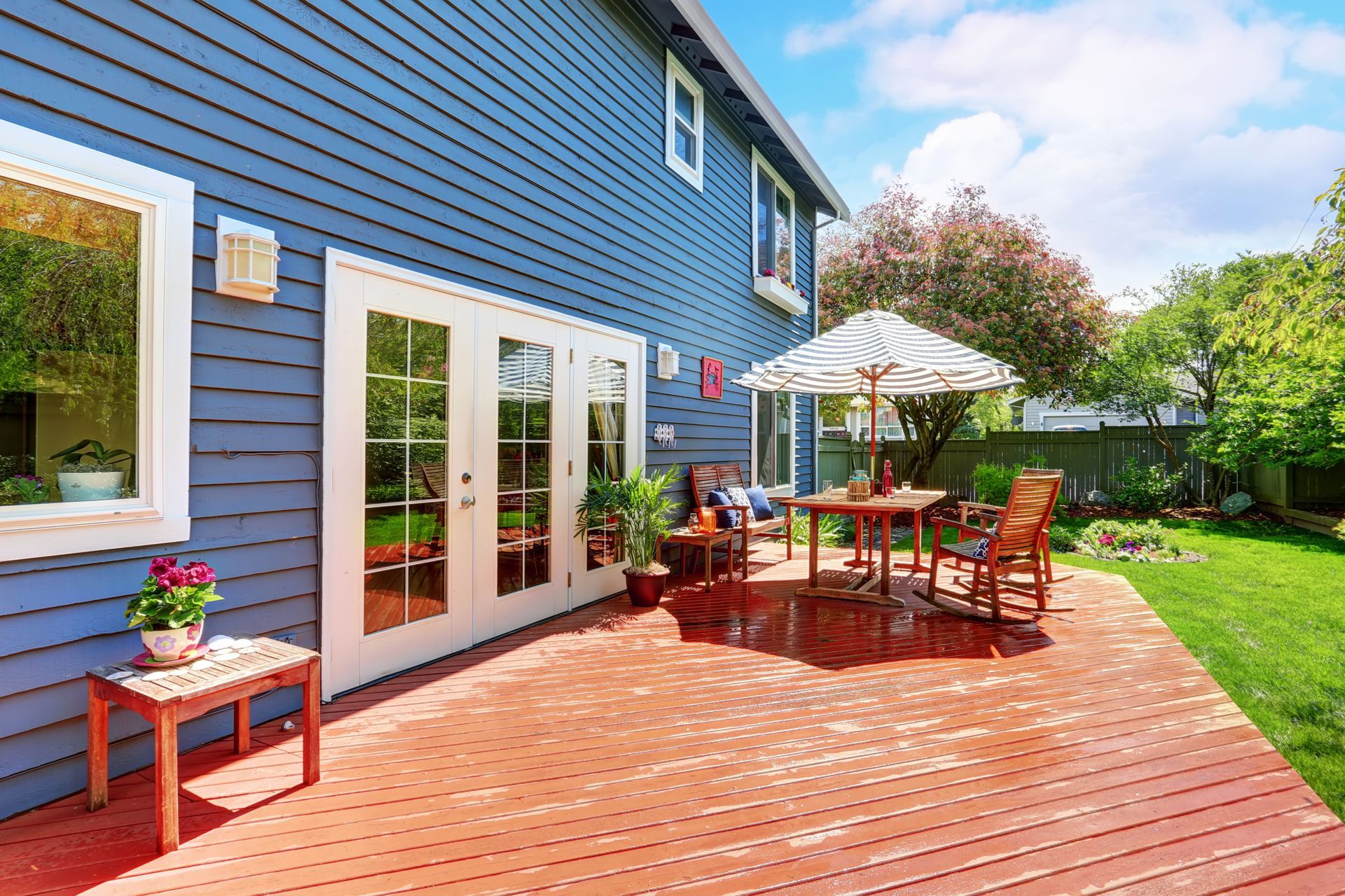 Blue house with white French doors opening to a red wooden deck and patio table with umbrella in a sunny backyard