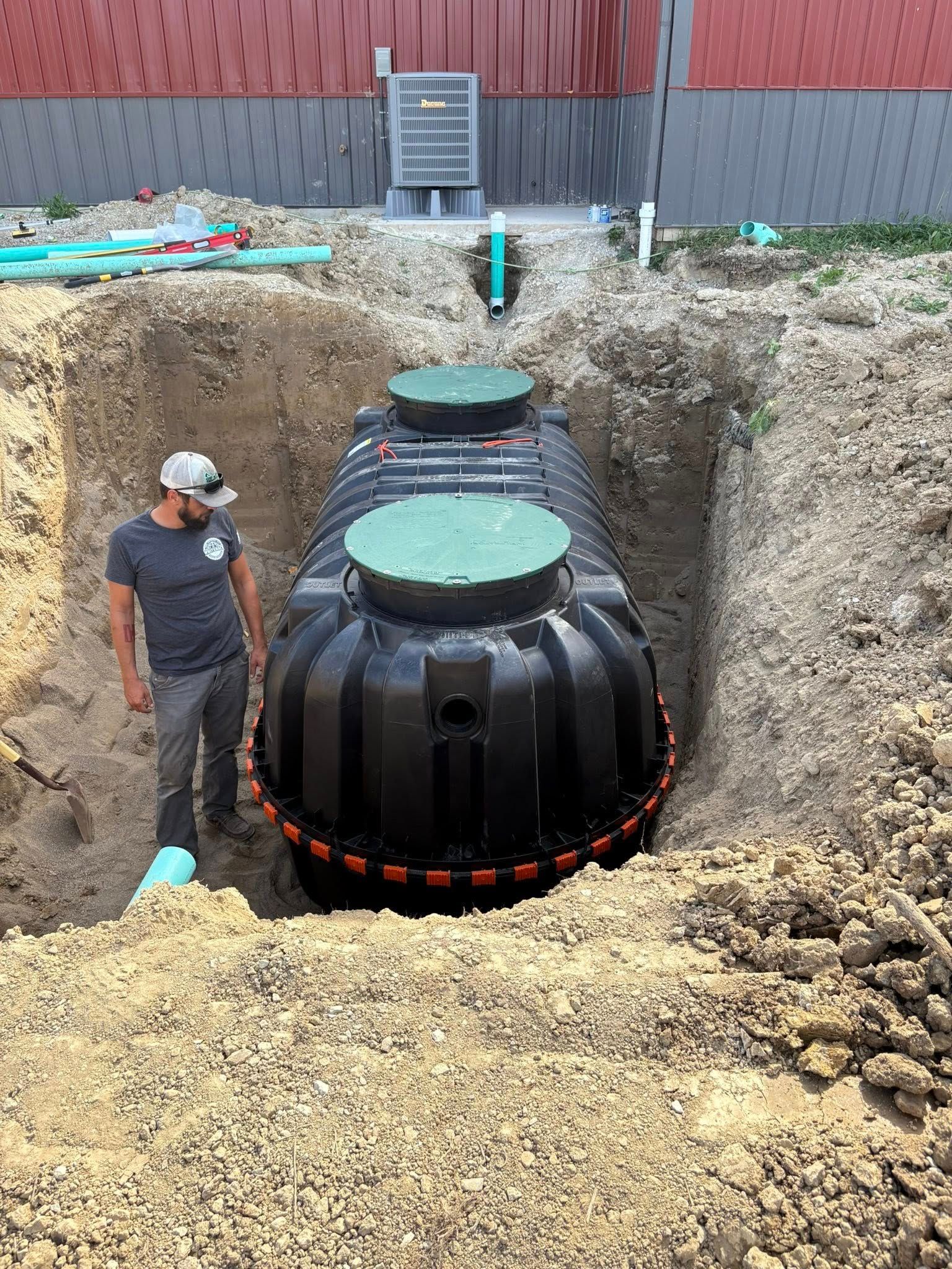 Two black underground tanks in a pit, with a worker standing beside them on a construction site.