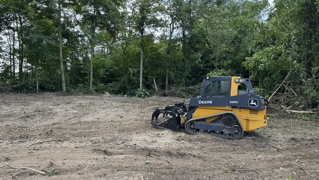Yellow tracked bulldozer clearing a dirt lot beside a wooded tree line