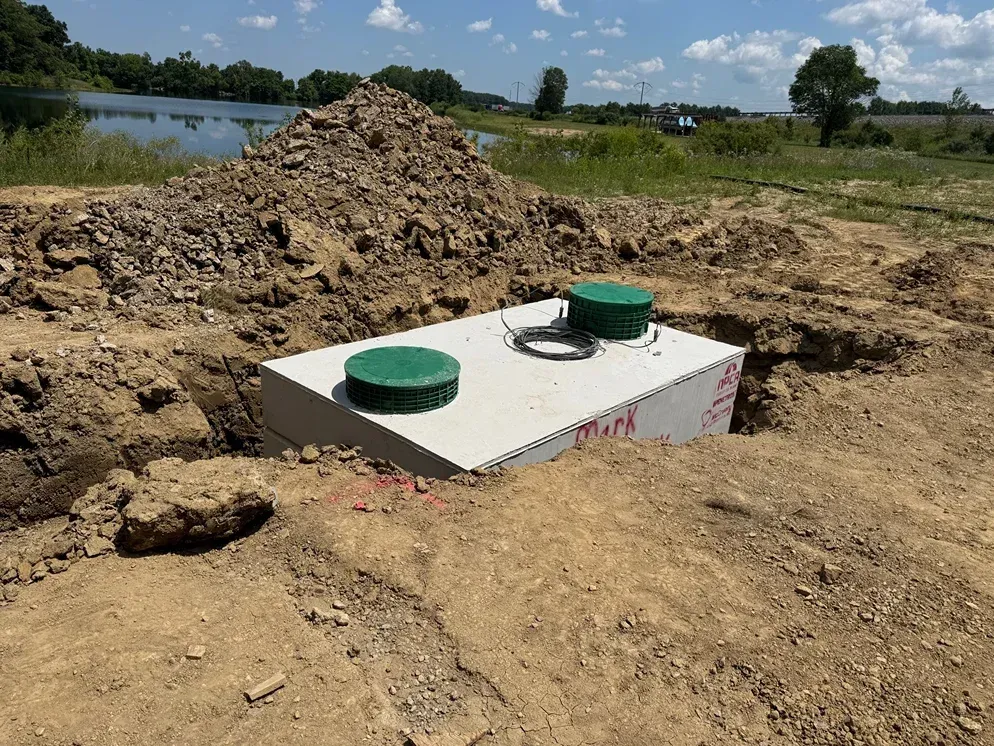 Rectangular underground utility tank with two green caps in a dirt excavation near water under a cloudy sky