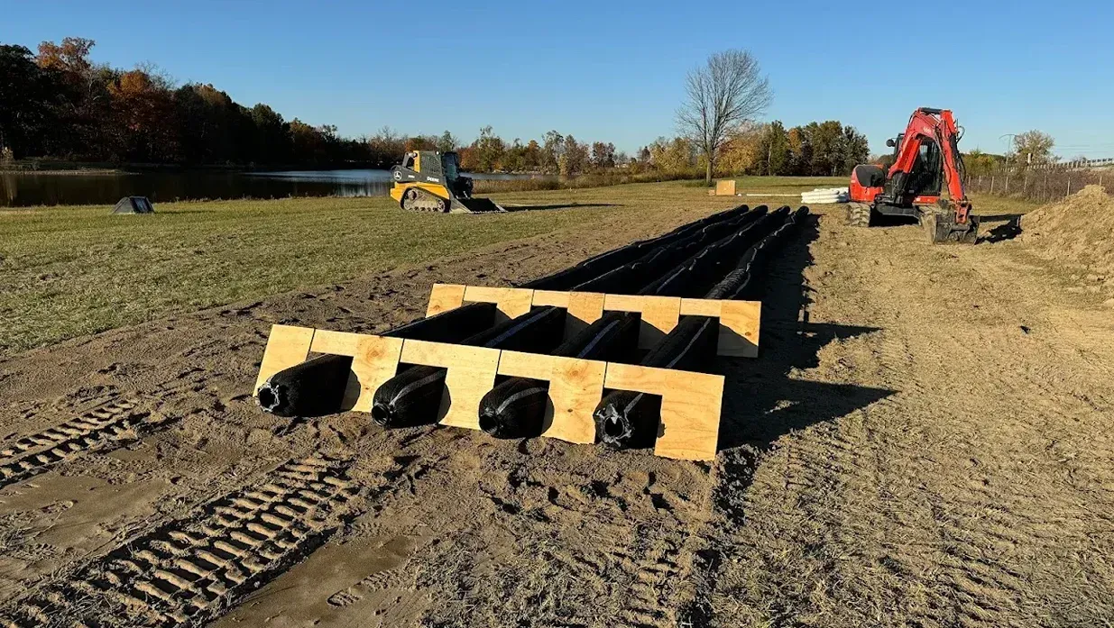 Long black drainage pipe sections on a dirt field, with yellow and red excavators in the background