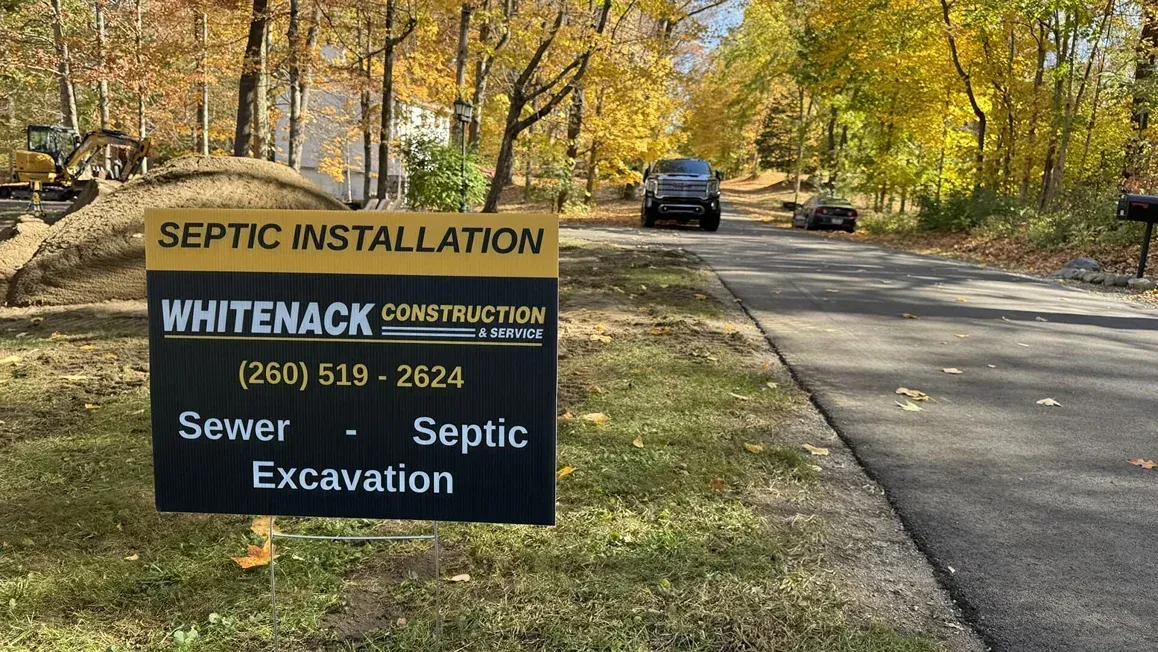 Septic installation sign for Whiteback Construction beside a wooded roadside with a vehicle in the distance