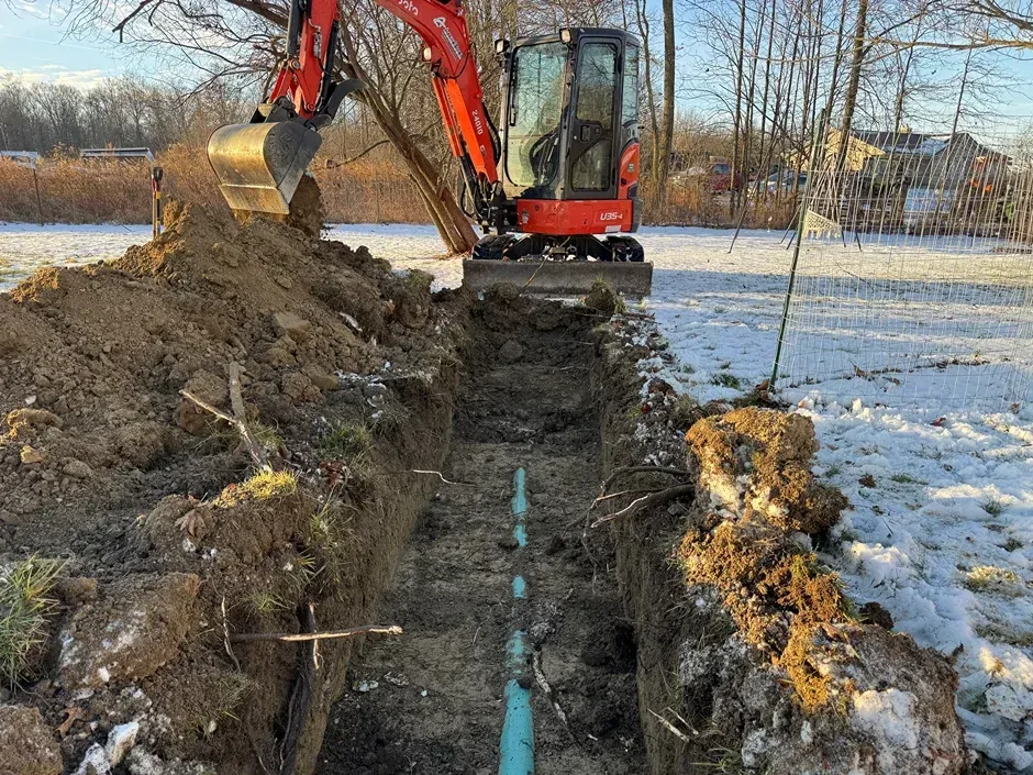 Excavator digging a trench through snowy ground beside piled soil in a field