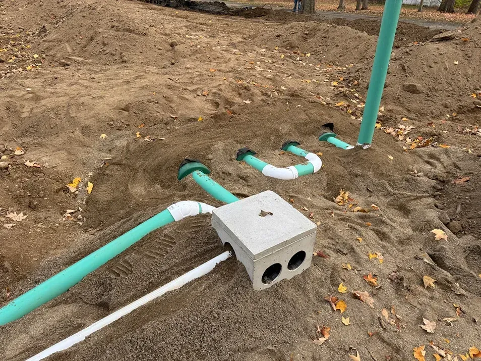 Green conduit pipes in a dirt trench leading to a gray utility box with two circular openings.