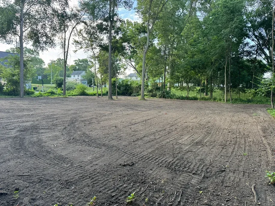 Freshly graded dirt clearing with trees and houses in the background under a cloudy sky