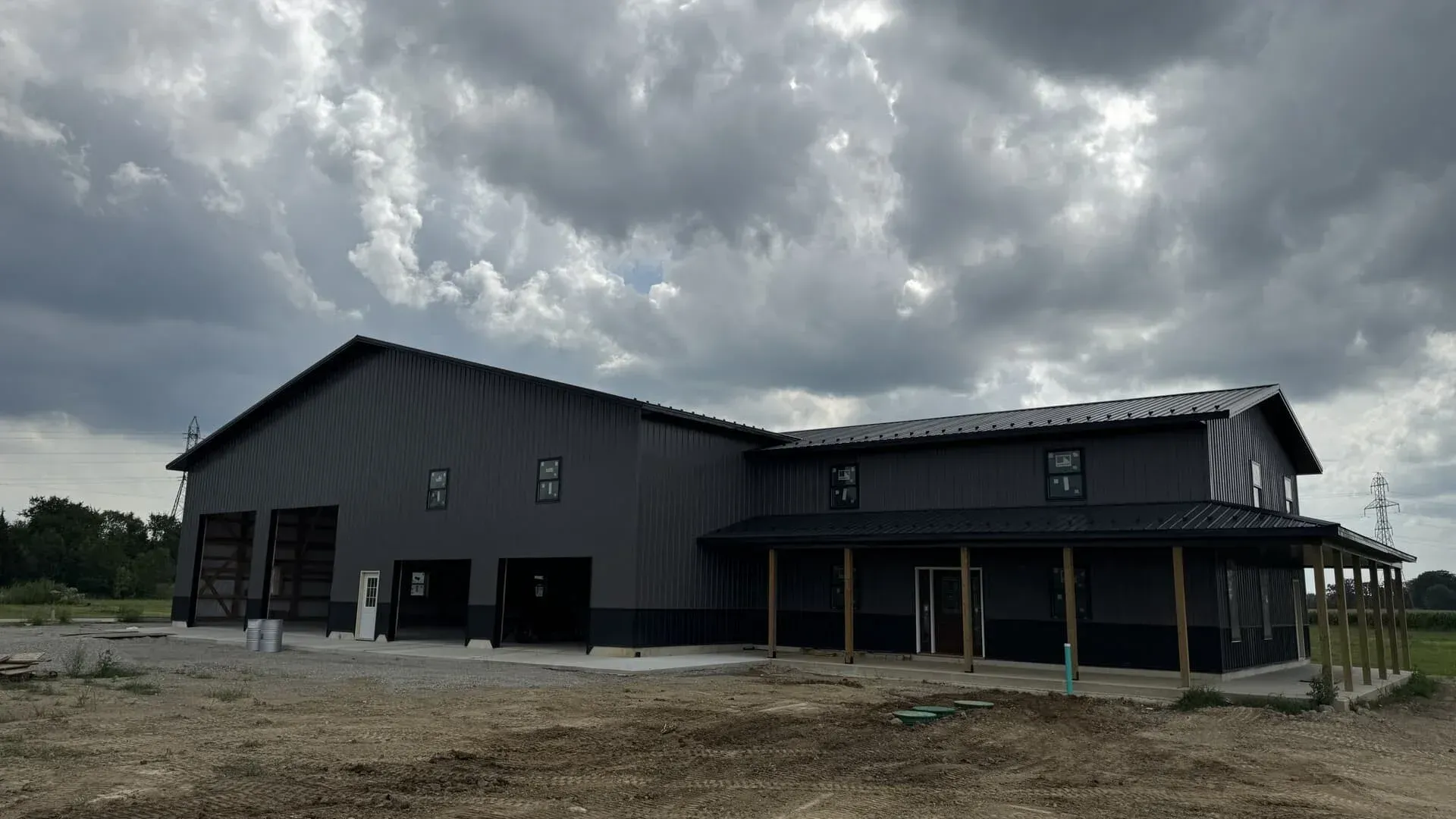 Large dark gray barn-style building under construction on a gravel lot beneath cloudy skies
