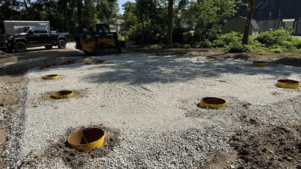 Gravel lot with several rusted pipe openings and construction vehicles in the background under trees