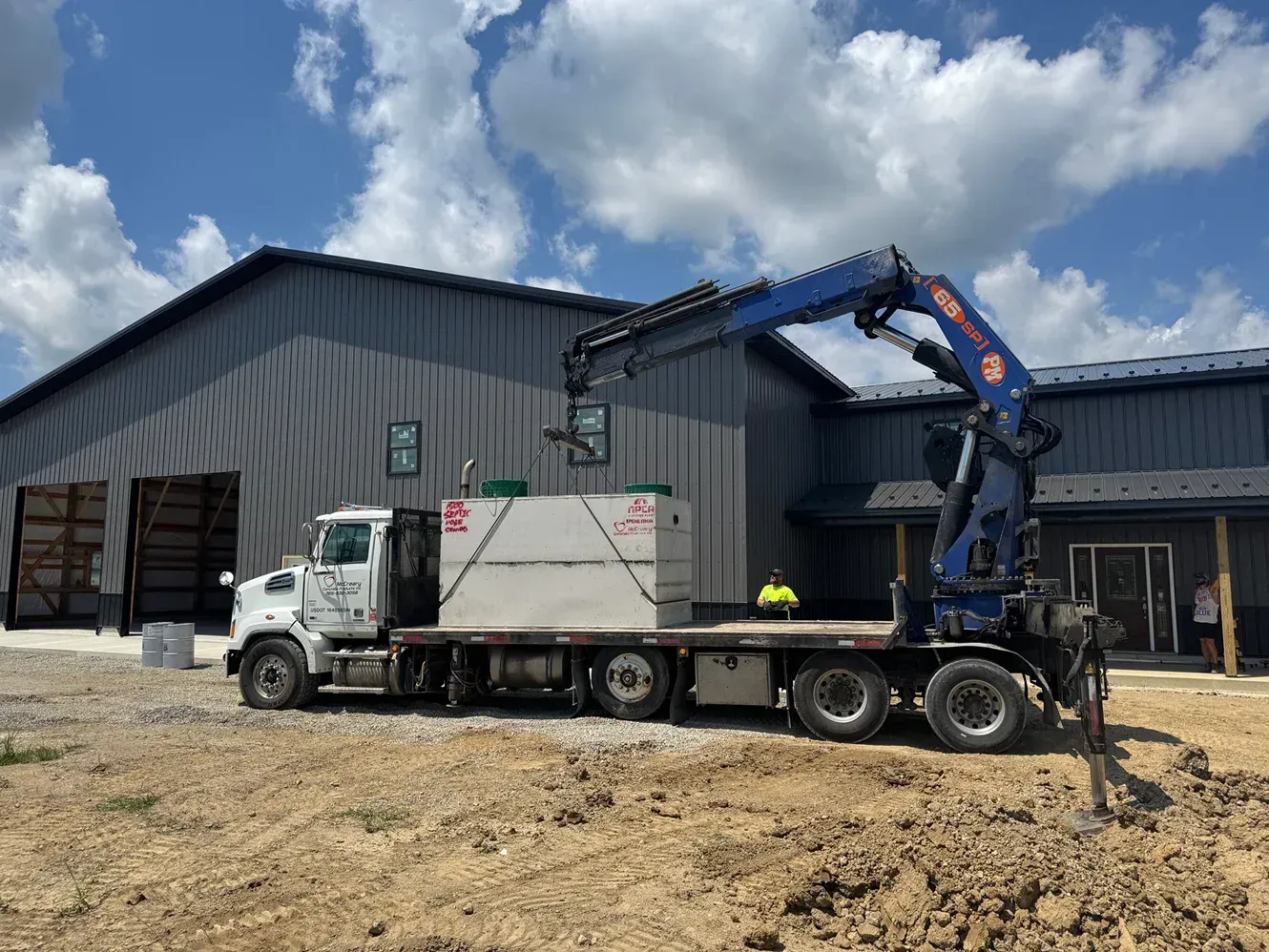 White flatbed truck with crane loading a container outside a metal warehouse under a cloudy sky