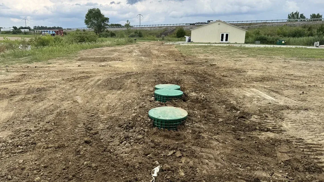 Rural dirt path with two green manhole covers leading toward a small building in a grassy field.
