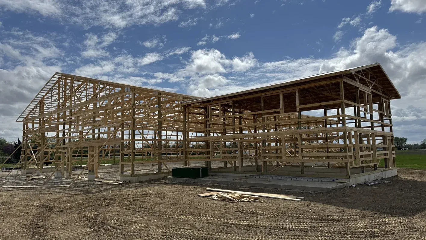 Wood-framed building under construction on a gravel lot beneath a cloudy blue sky