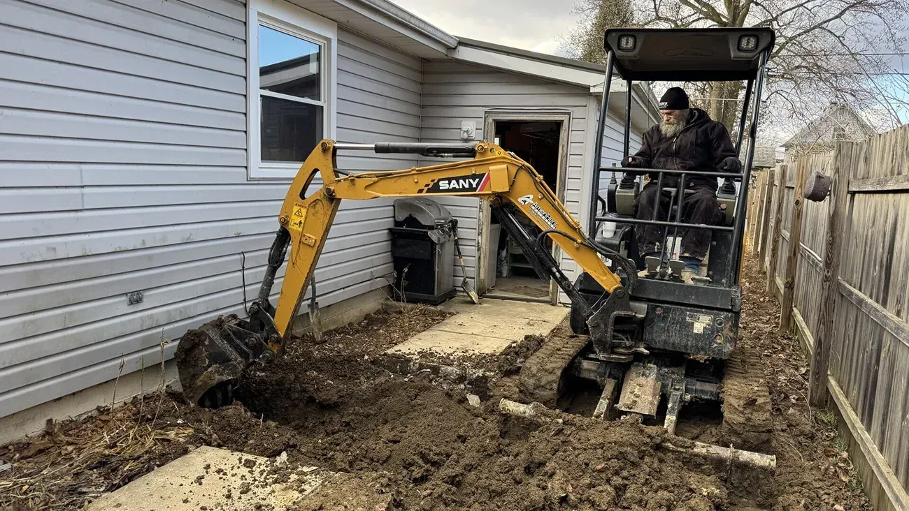 Yellow mini excavator digging along a house foundation beside a white-sided home.