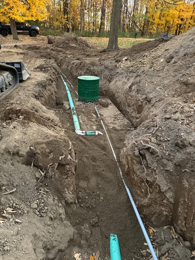 Trenched utility line with green pipe and sewer access in a dirt excavation, autumn trees in background