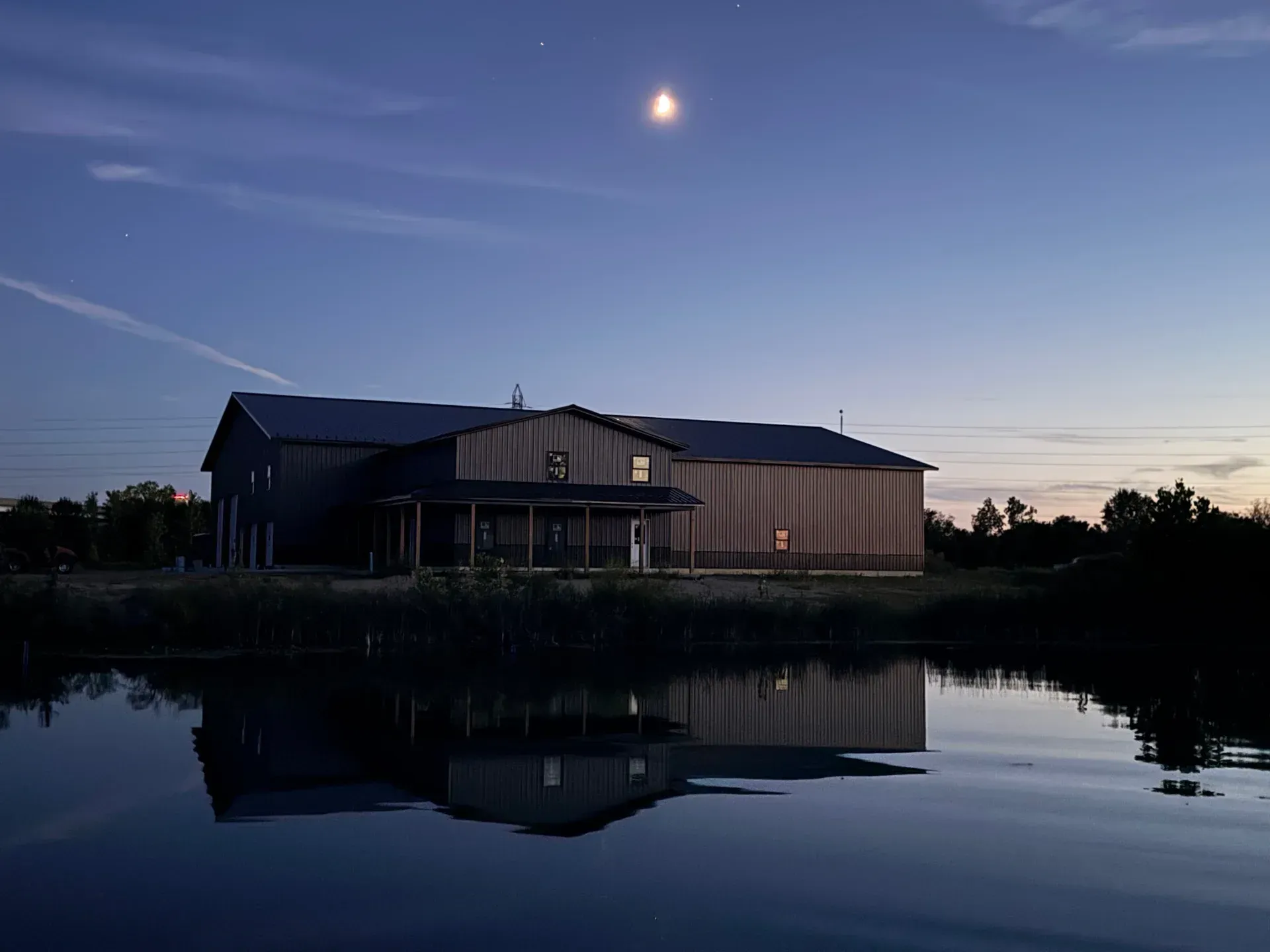 Modern house by calm water at dusk, with a bright moon and reflection in the foreground.