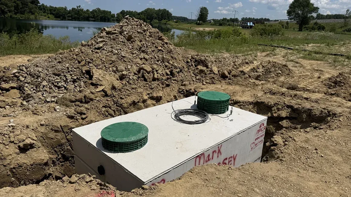 Rectangular concrete utility tank with two green caps, partly buried in a dirt mound near a field and water.