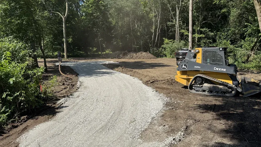 Small bulldozer grading a gravel path through a wooded area in daylight