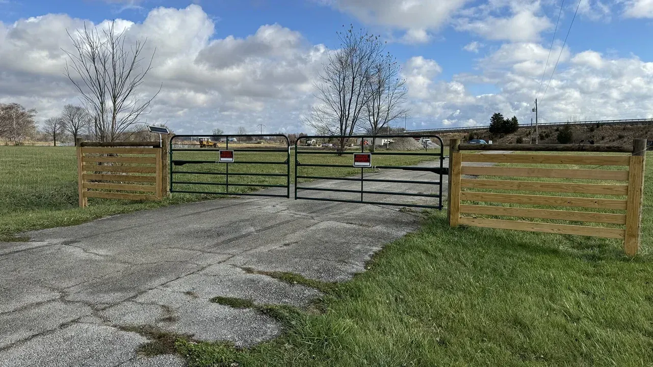 Wooden farm gate beside a cracked concrete driveway opening onto a grassy field under a cloudy sky