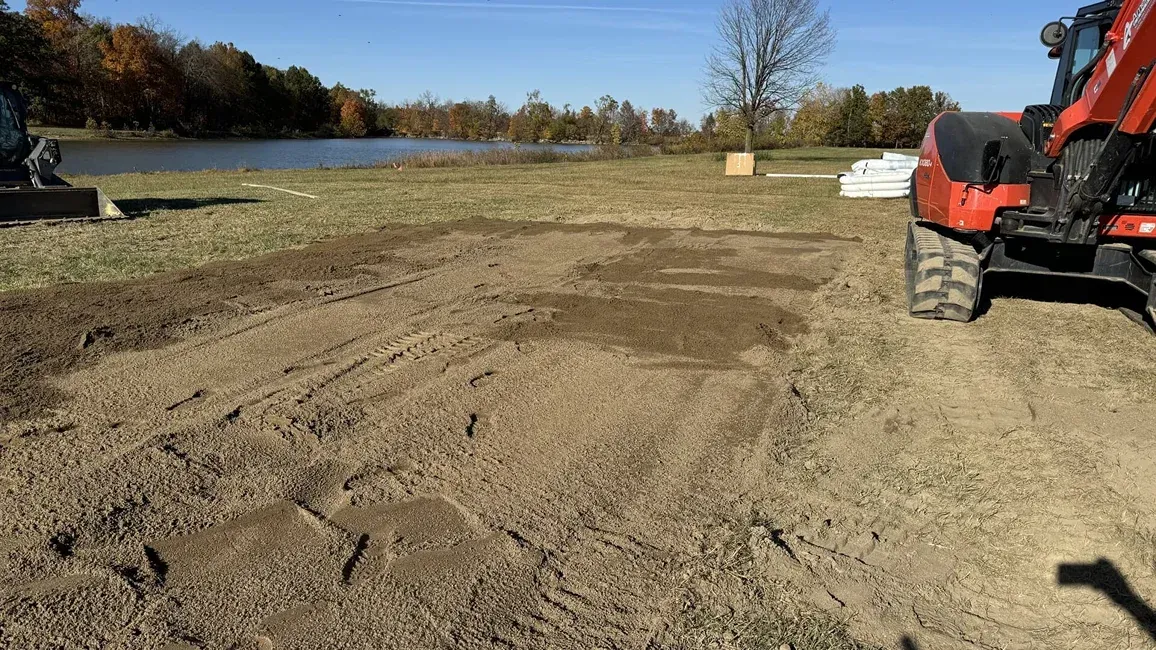 Dirt construction site beside a lake, with an orange excavator on the right under a clear blue sky.