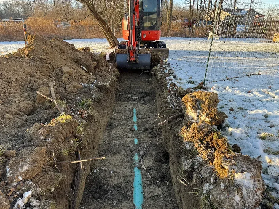 Excavator digging a trench through snowy ground beside a frozen field