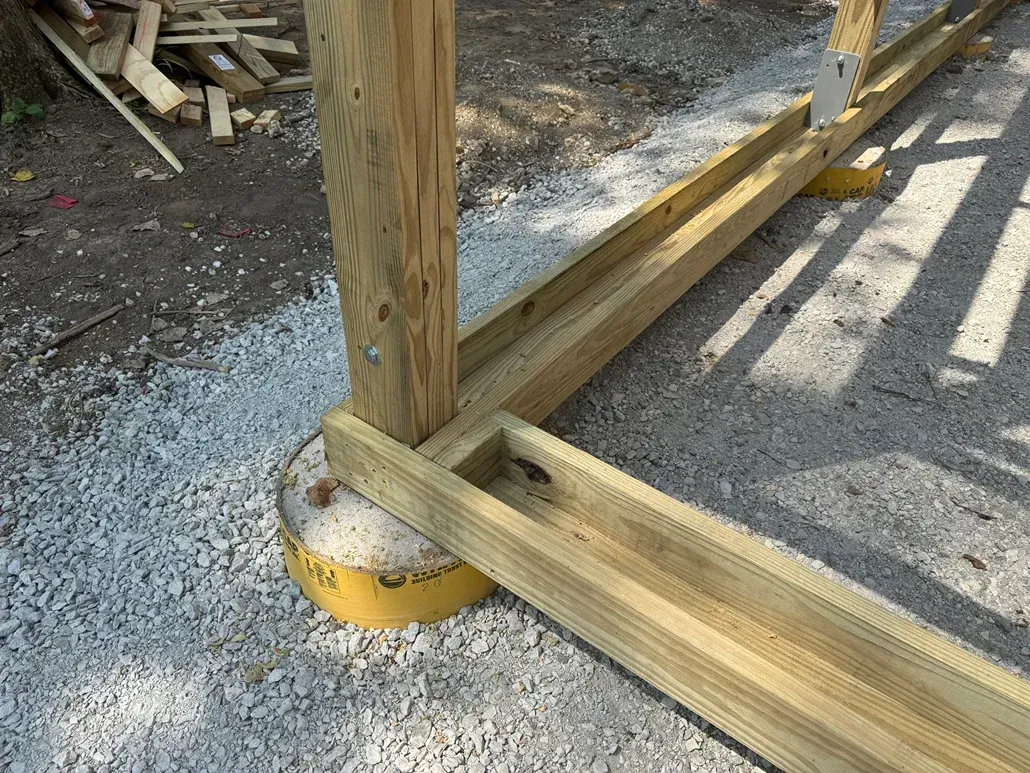 Wooden deck framing on a yellow footing base over gravel at a construction site