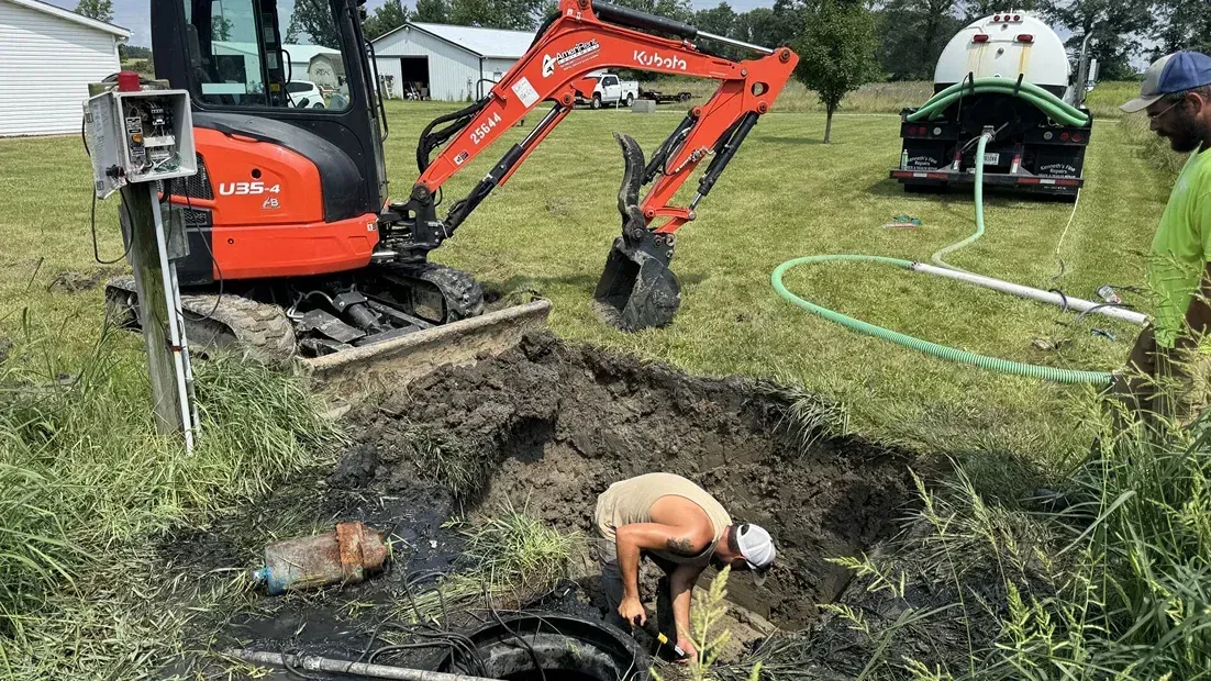 Orange excavator digging a muddy trench while a worker kneels in a yard