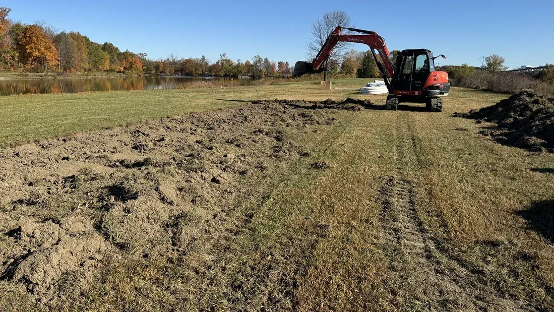Excavator grading a dirt field beside a grassy edge under a clear sky