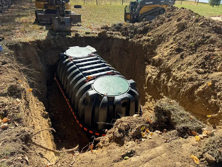 Excavator lowers a large black cylindrical tank into a deep dirt trench at a construction site.