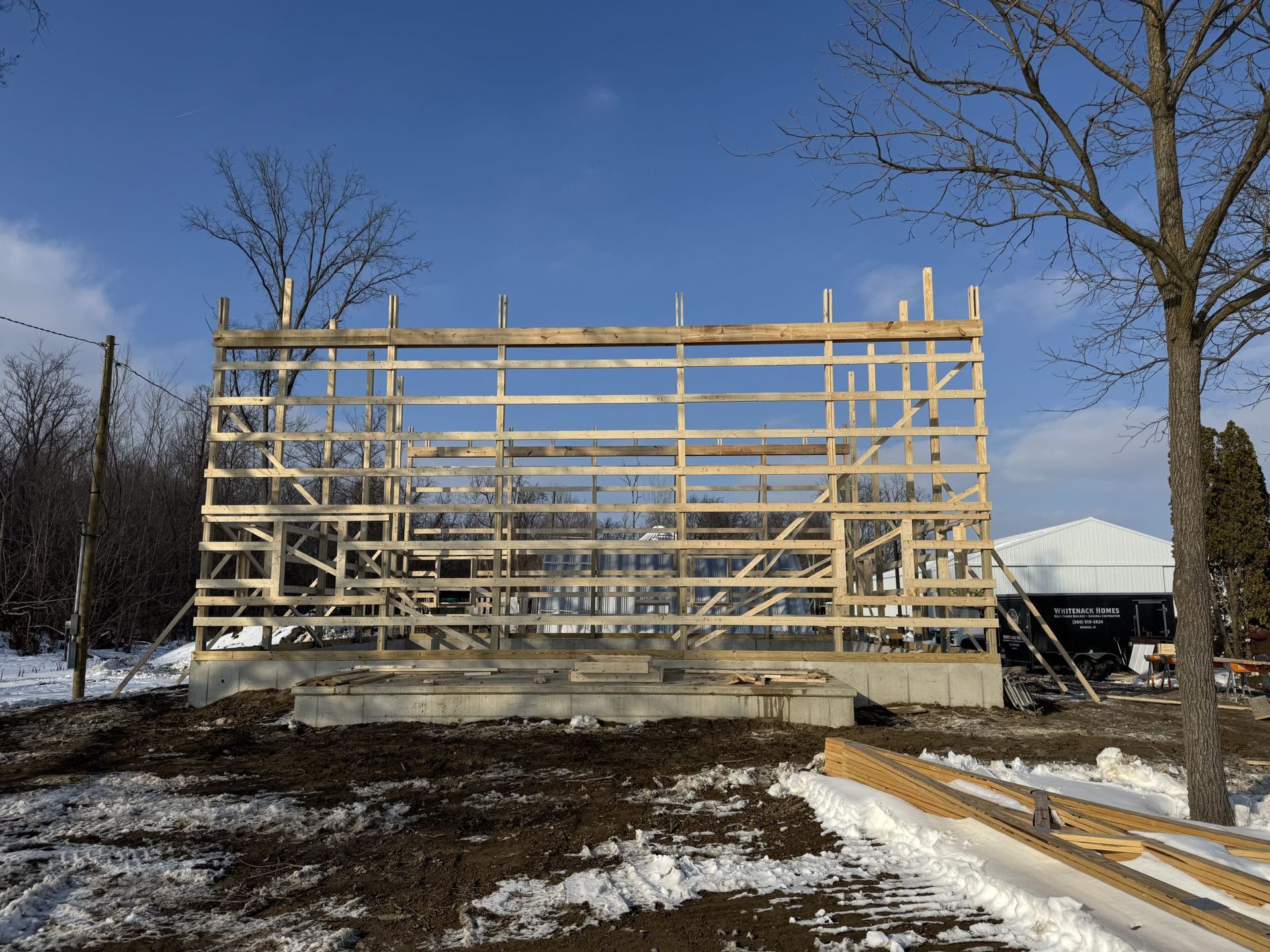Wood-framed building under construction on a snowy site with bare trees and a blue sky