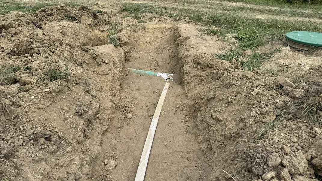 Trench in dry dirt with a white measuring tape laid along the bottom and a green lid in the background