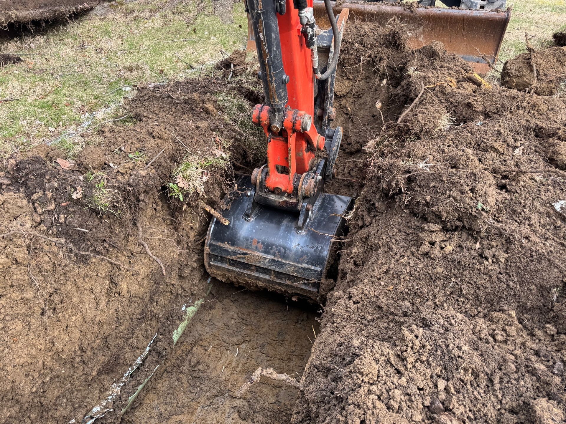 Excavator digging a narrow trench in muddy soil, with orange arm and bucket exposed.