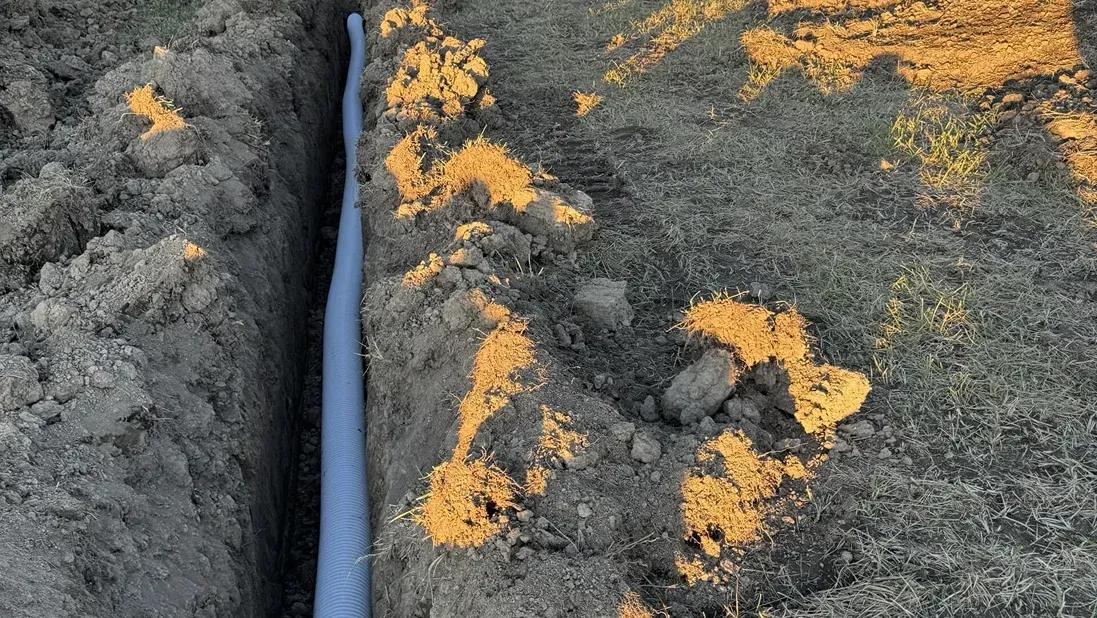 Black pipe between two mounds of gray soil with orange insulation or debris along the trench.