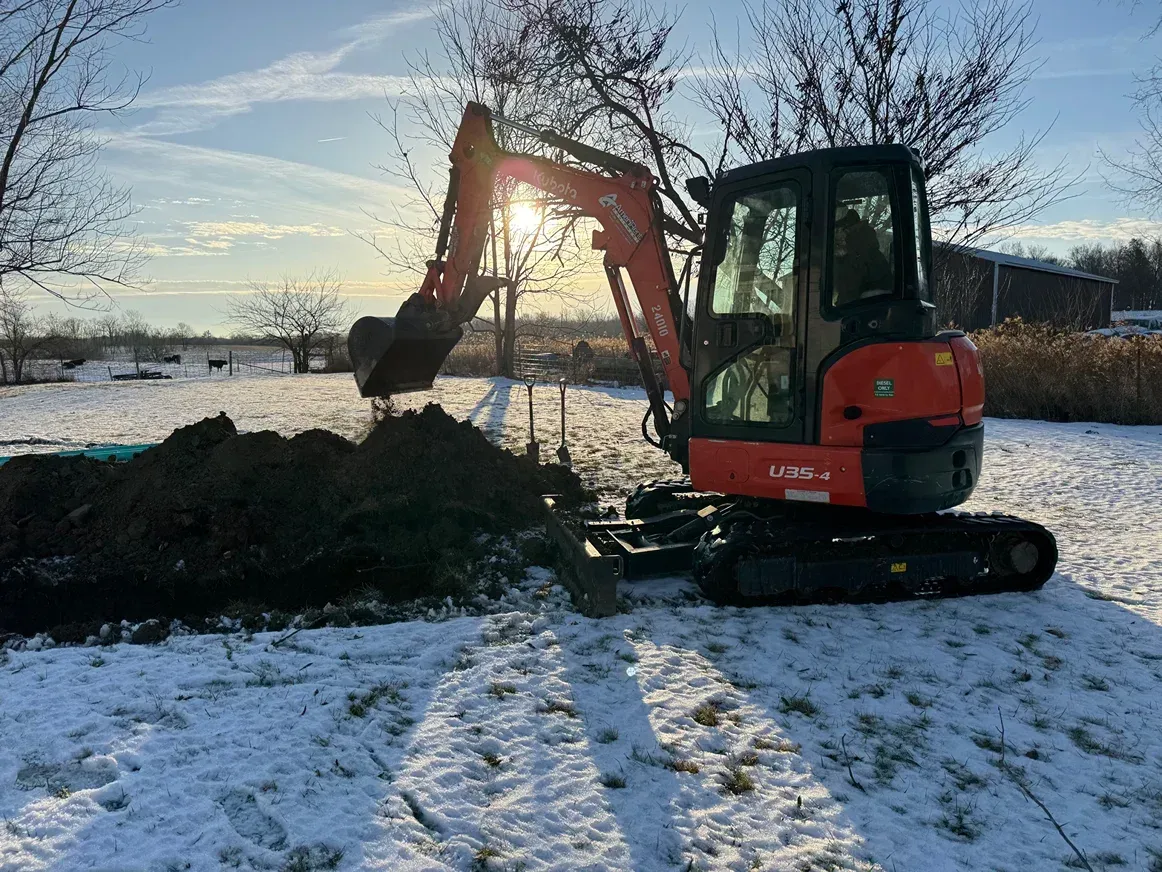 Orange excavator digging a dirt pile in a snowy field at sunrise