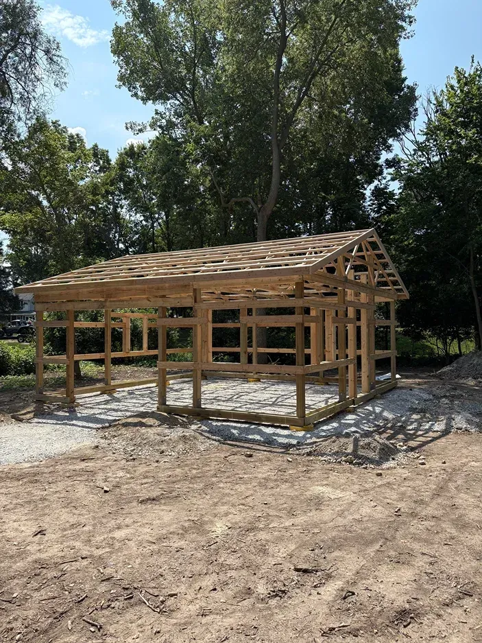 Wooden house frame under construction on a gravel lot, surrounded by trees and dirt.