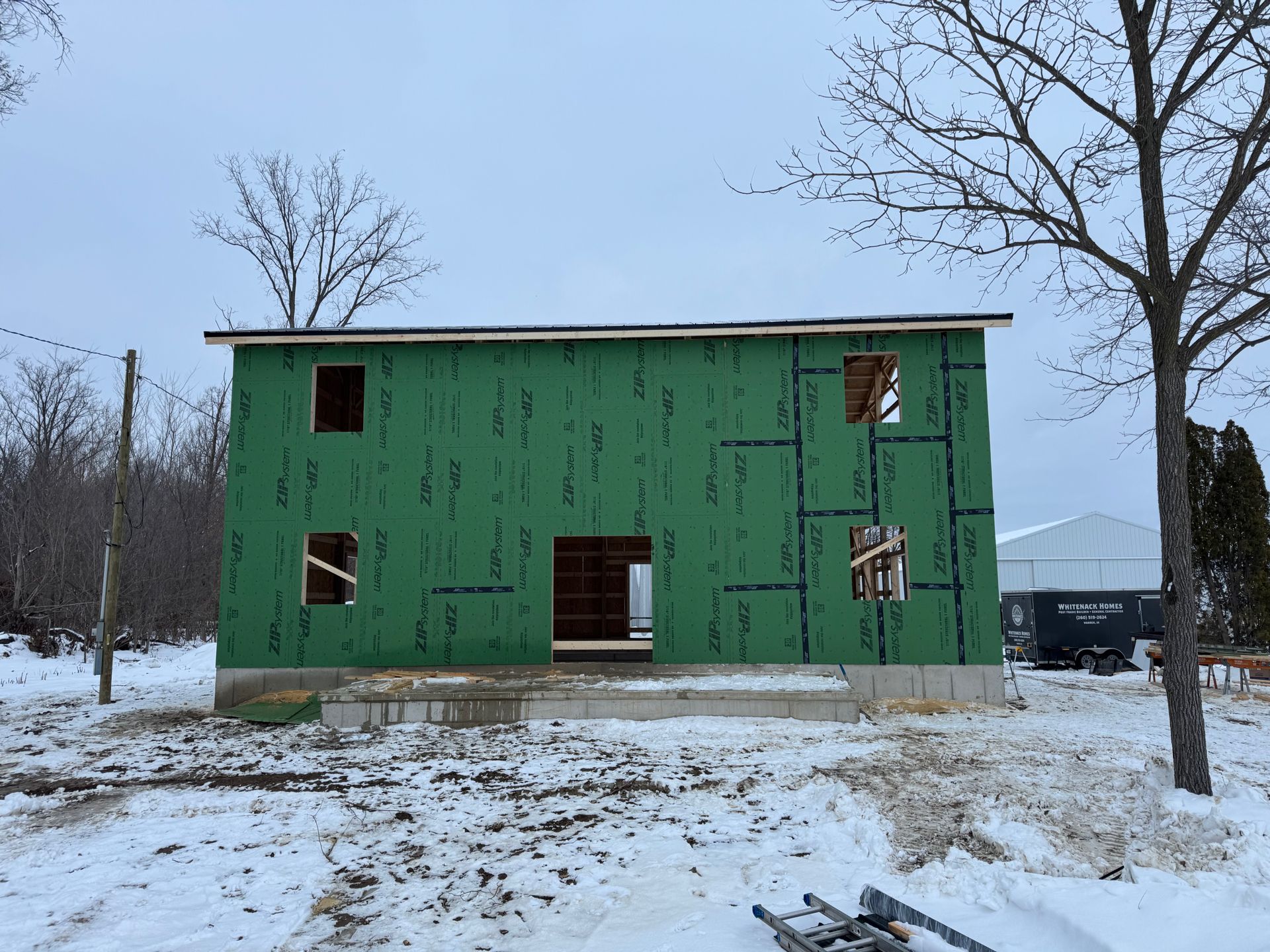 Green unfinished house in a snowy yard with bare trees and a cloudy winter sky