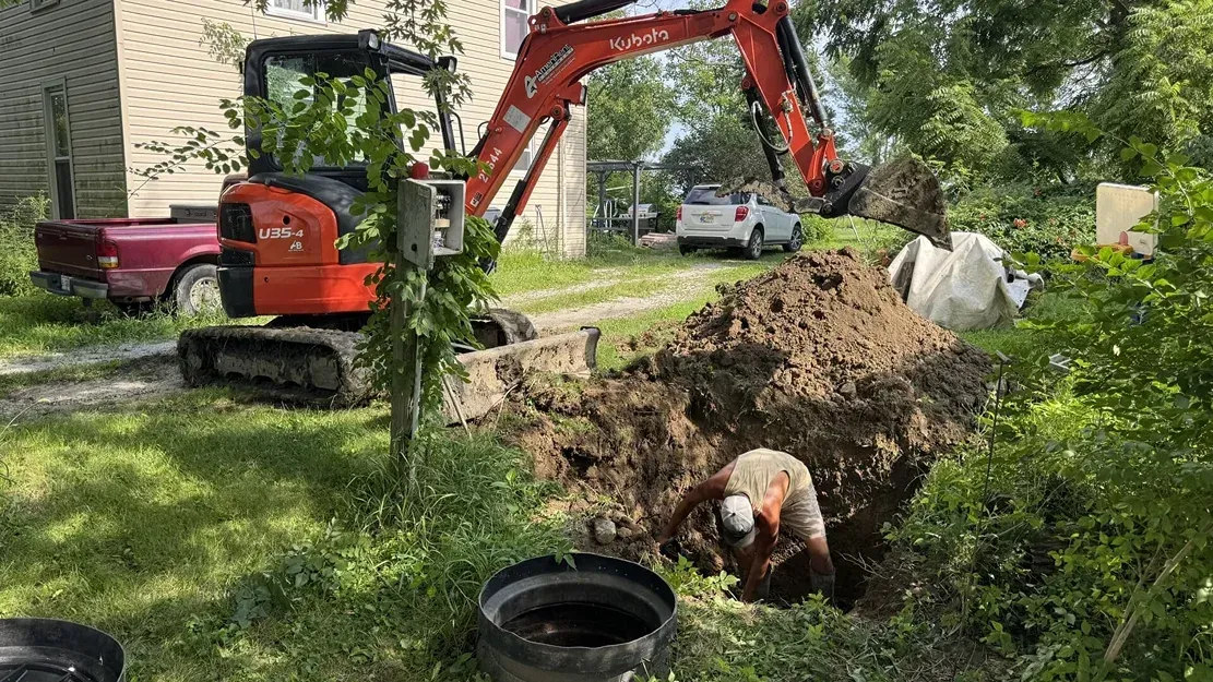 Orange excavator digging a trench in a leafy yard while a worker crouches in the dirt