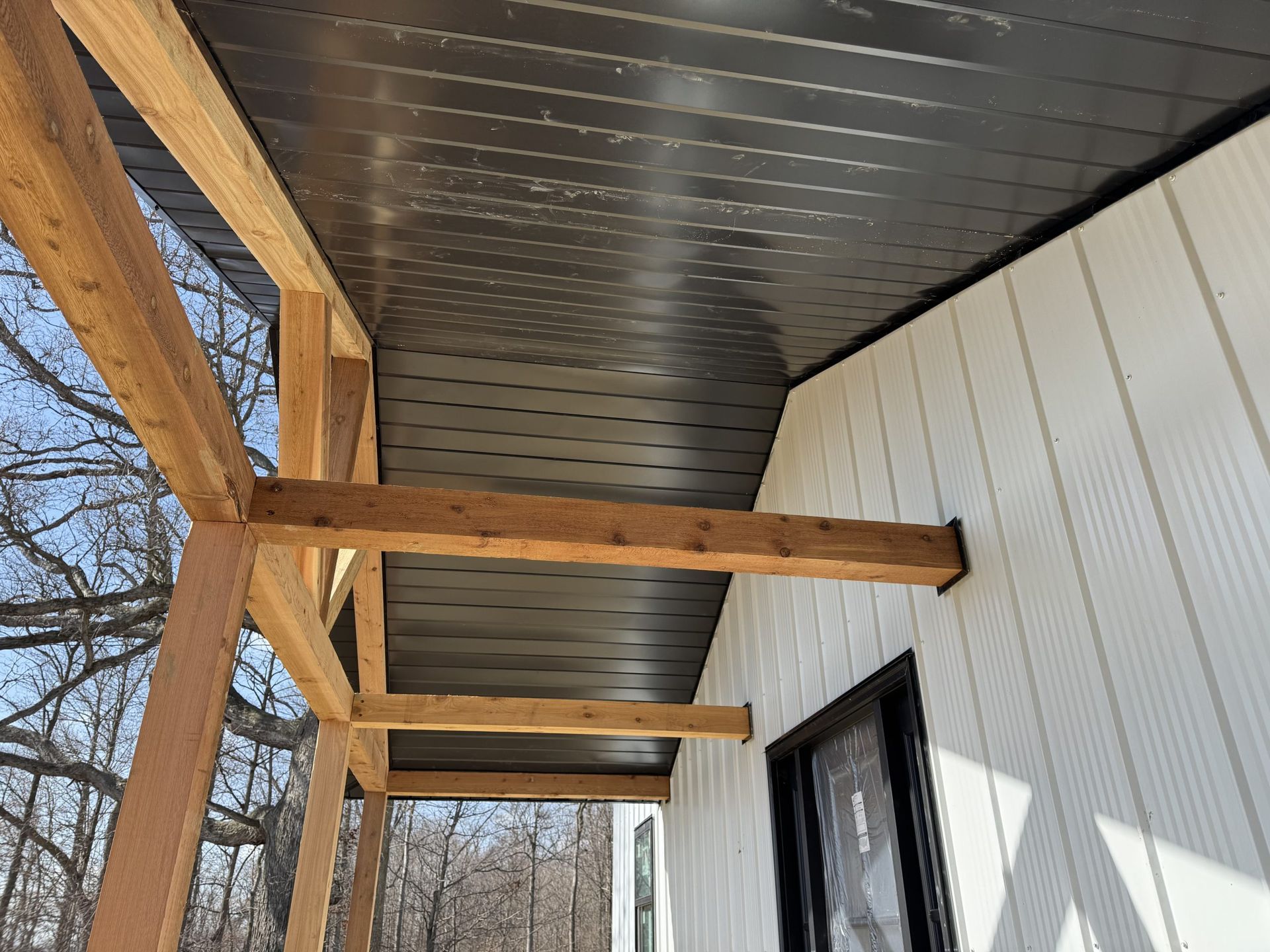 Covered porch with exposed wooden beams and white siding beside a tree-lined yard