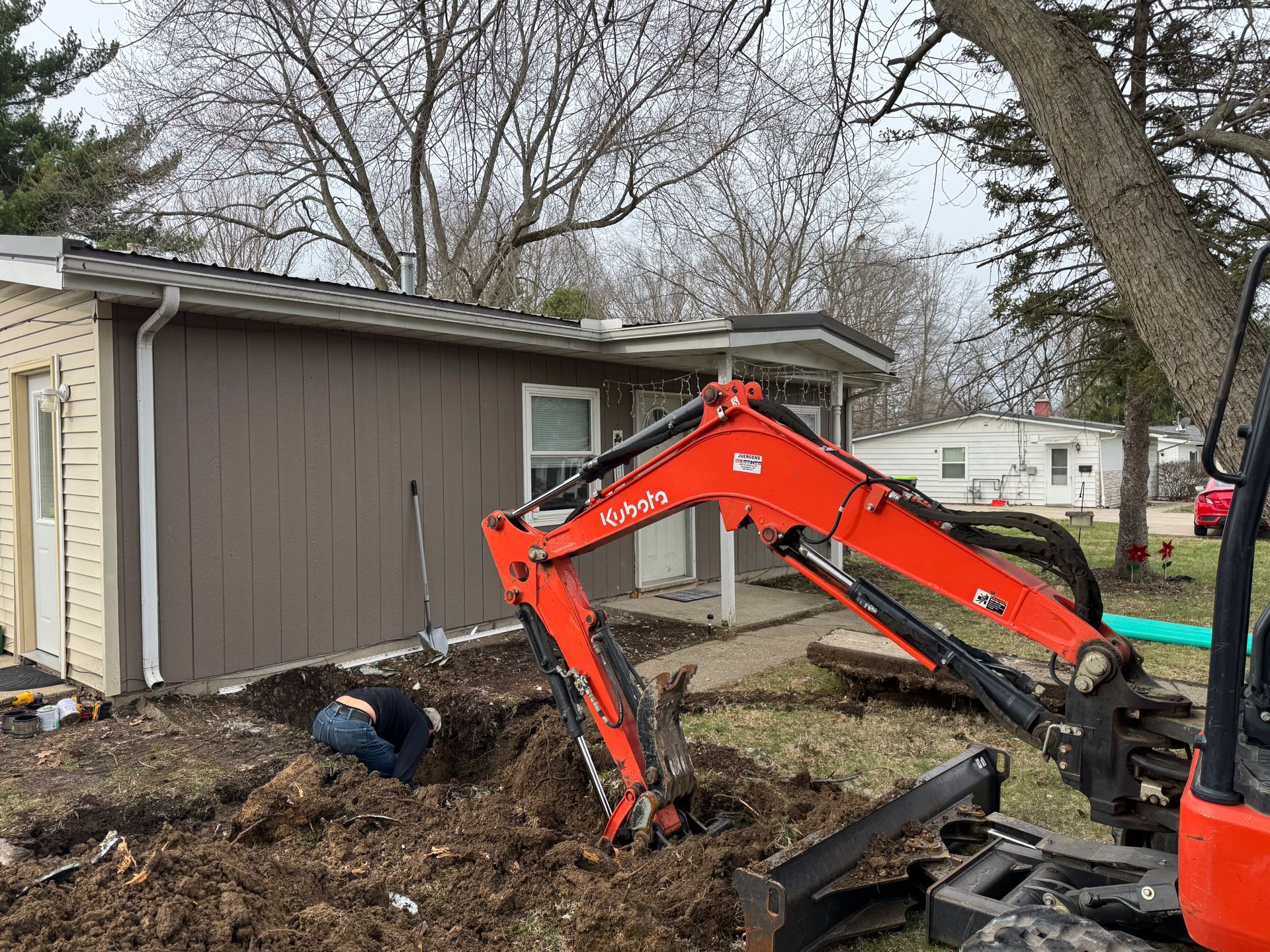 Orange excavator digging near a garage while a worker shovels dirt in a yard