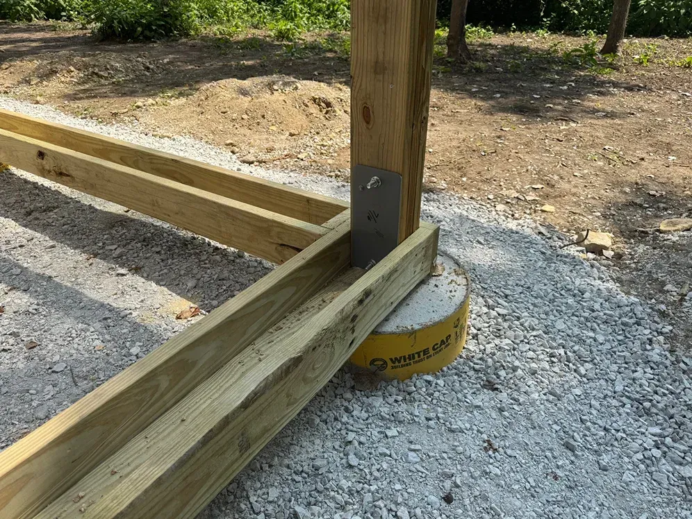 Wooden deck frame secured to a vertical post on a gravel surface, with a metal base plate.