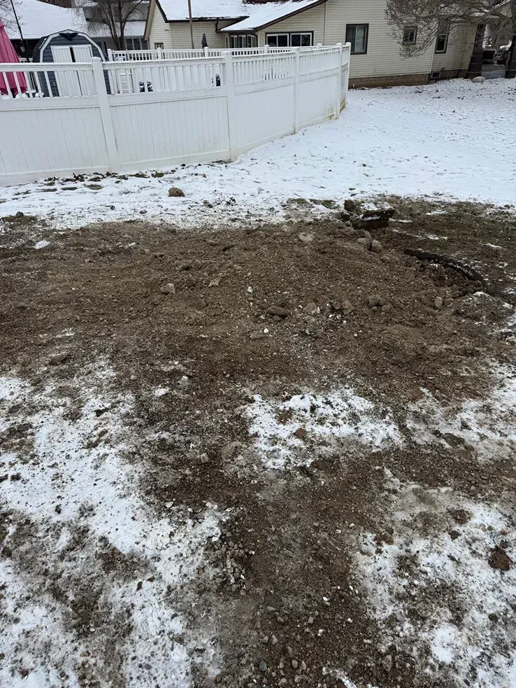 Snowy backyard with a large muddy patch and a white fence in the background