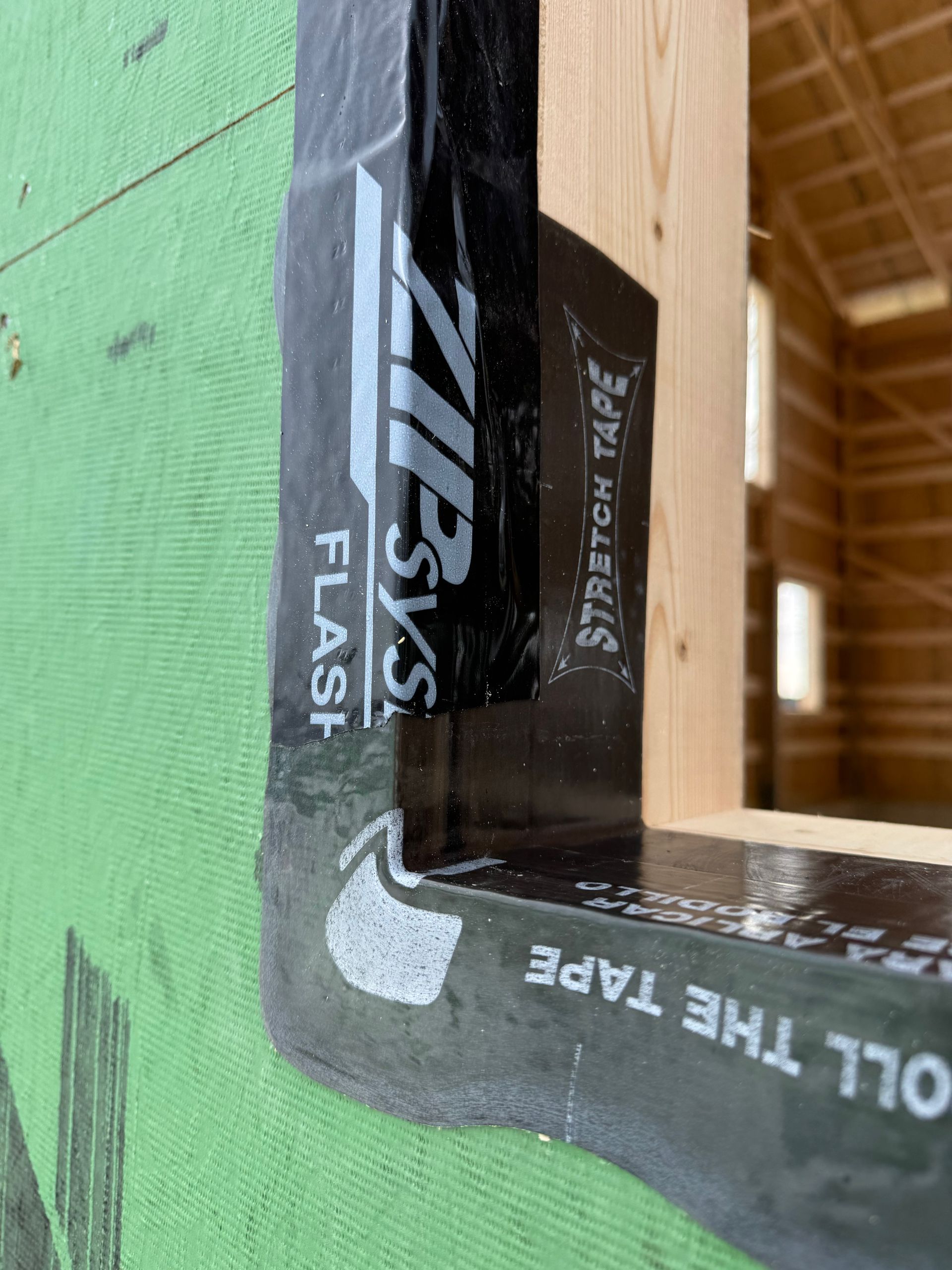Close-up of a wooden frame joint wrapped with black flashing tape on green sheathing.