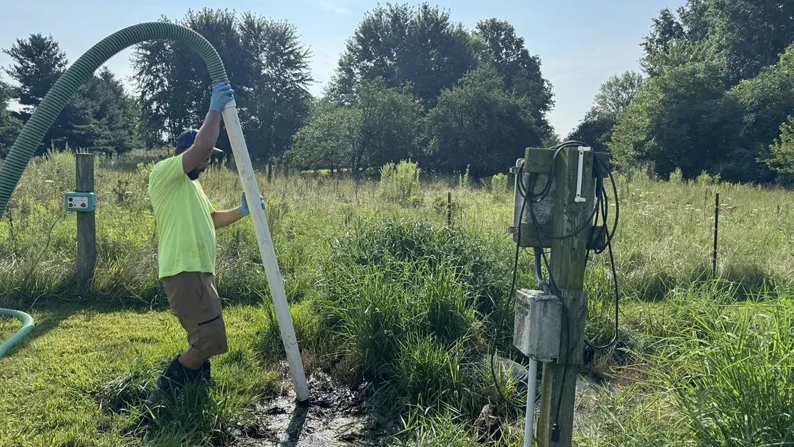 Person in neon shirt working a hand pump in a sunny grassy field with trees in the background