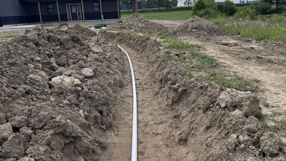 A narrow pipe runs through a freshly dug trench beside a building and dirt piles.